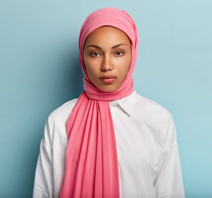 arabian-woman-with-calm-serious-expression-has-dark-skin-wrapped-pink-veil-wears-white-shirt-has-no-make-up-natural-beauty-models-blue-wall-close-up-shot-muslim-lady