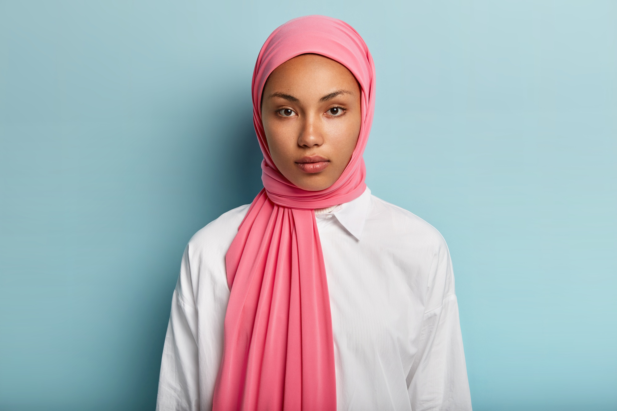 arabian-woman-with-calm-serious-expression-has-dark-skin-wrapped-pink-veil-wears-white-shirt-has-no-make-up-natural-beauty-models-blue-wall-close-up-shot-muslim-lady